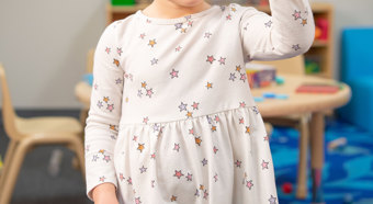 Una niña pequeña de tez clara y rubia con el pelo atado, con un vestido claro, sonriéndo y mostrándole los cinco dedos de su mano extendida a la cámara.