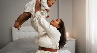 Una mujer adulta sostiene a un bebé en el aire, ambos se miran y sonríen. La escena transcurre en un ambiente hogareño y acogedor.