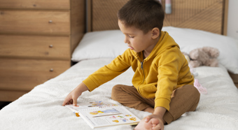 Un niño pequeño que viste una camisa amarilla de manga larga se sienta en la cama mientras lee un libro infantil.