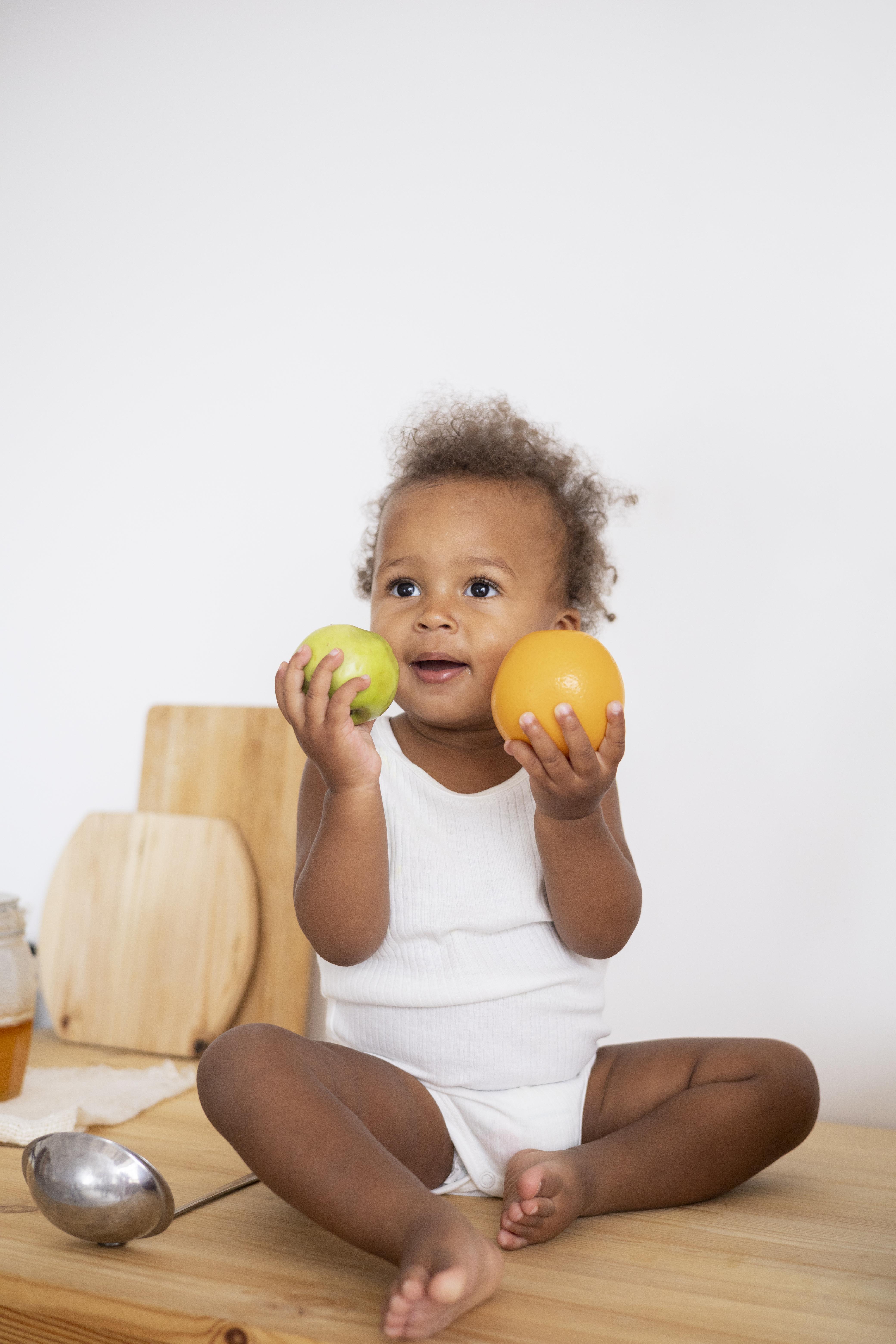 Un niño pequeño sentado en la encimera de la cocina, vestido todo de blanco, sostiene dos frutas en las manos mientras sonríe y mira a lo lejos.