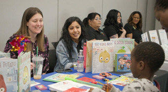 Dos mujeres sentadas en un stand en un evento de proyección sonríen mientras hablan con un niño pequeño en el stand.