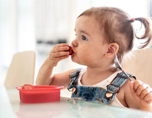 Una niña pequeña de tez clara, pelo rubio con coletas, con una remera blanca y un enterito de denim, sentada frente a una mesa con un tapper con fresas, comiendo una fresa con una mano y soteniendo una cuchara con la otra.