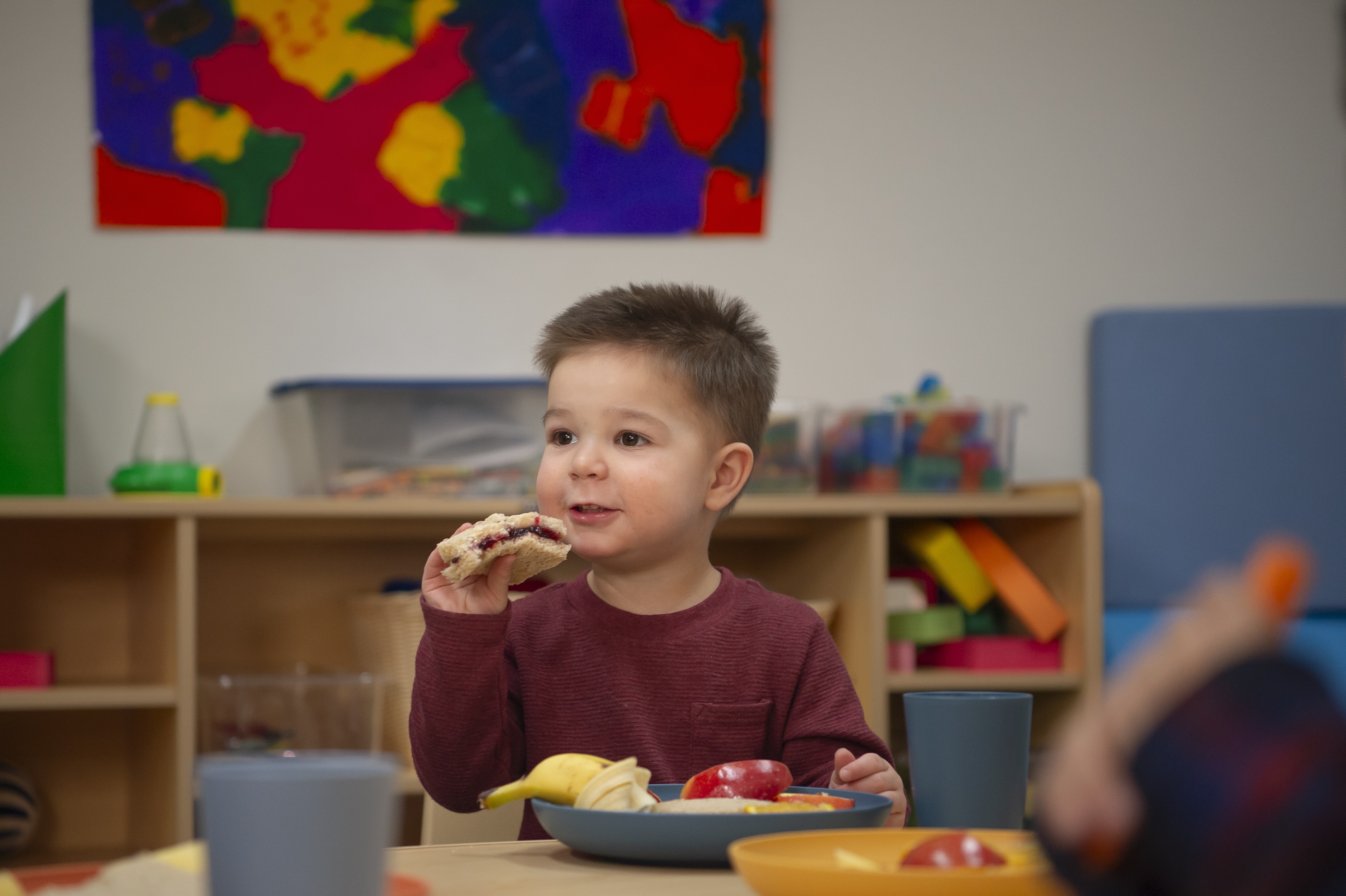 Un niño pequeño con tez clara y pelo castaño, sentado a la mesa comiendo un sandwich.