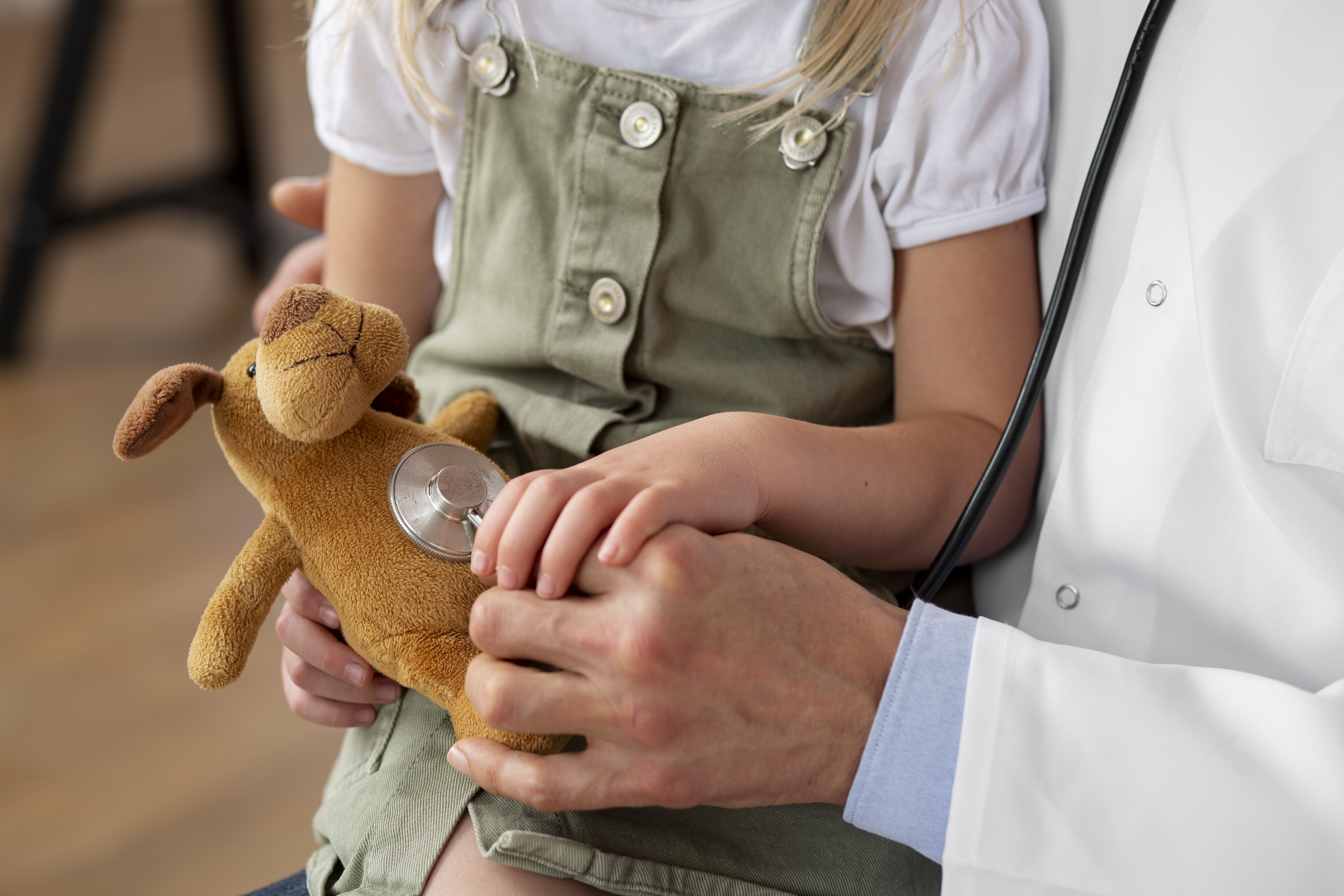 Primer plano de un profesional médico con bata blanca de laboratorio y una niña en su regazo, sosteniéndole la mano. La niña sostiene un animal de peluche y lleva un traje verde oscuro.