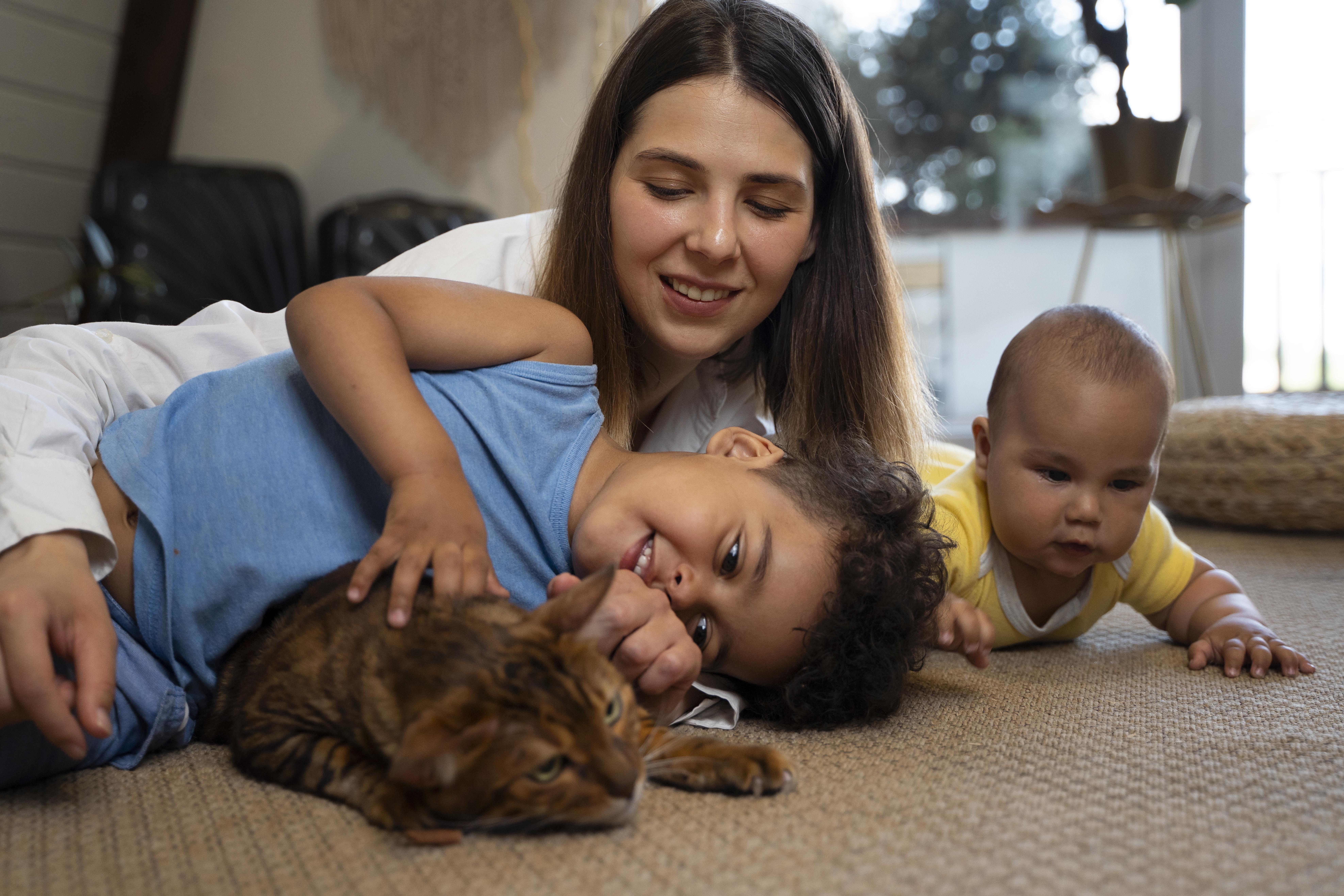 Un bebé y un niño pequeño tumbados en un suelo alfombrado con una jovencita y un gato marrón. Sonriendo y jugando con el gato.