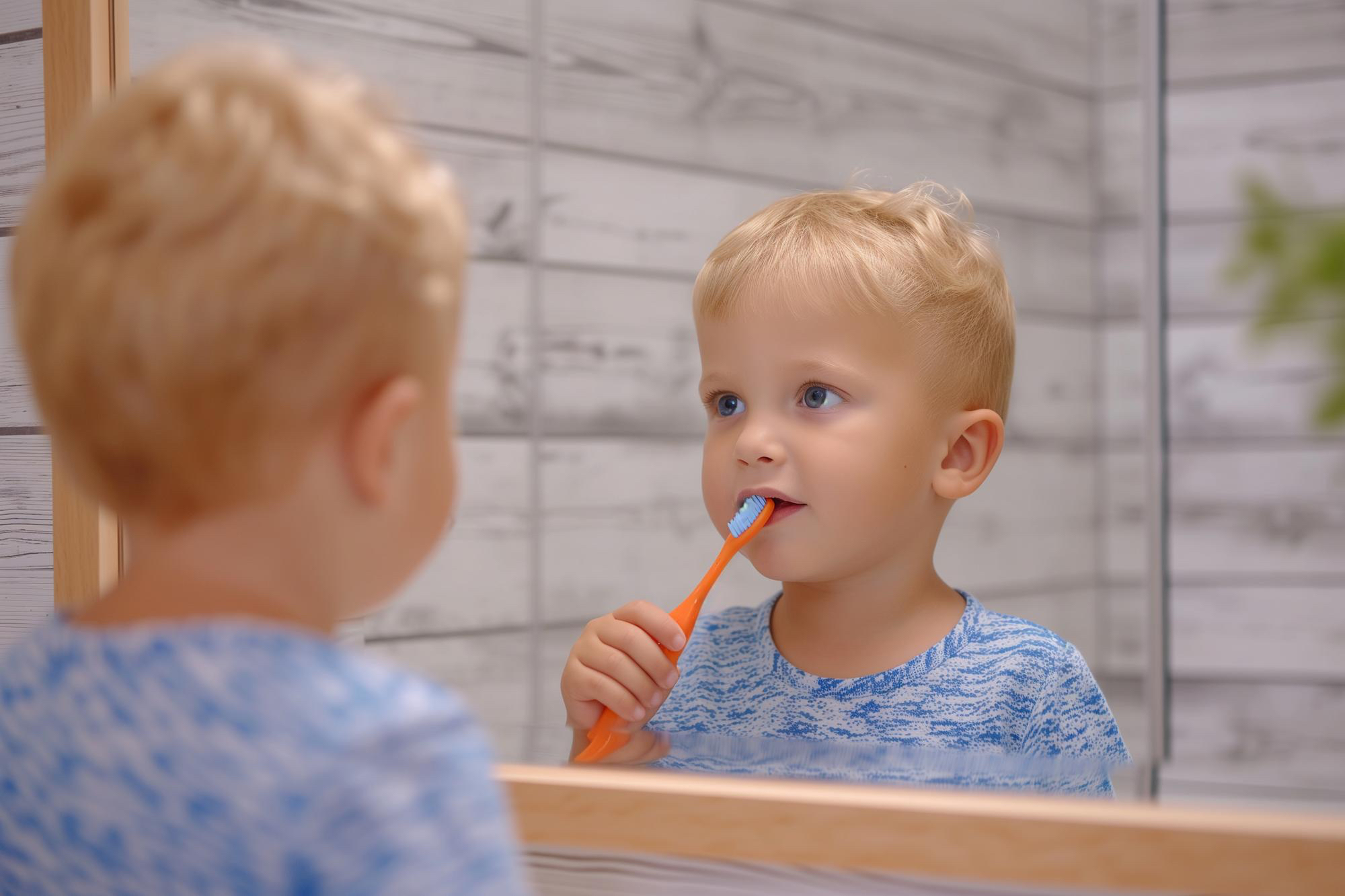 Un bebé con cabello rubio mira su reflejo en el espejo mientras se cepilla los dientes con un cepillo de dientes naranja