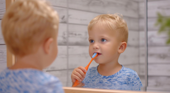 Un bebé con cabello rubio mira su reflejo en el espejo mientras se cepilla los dientes con un cepillo de dientes naranja