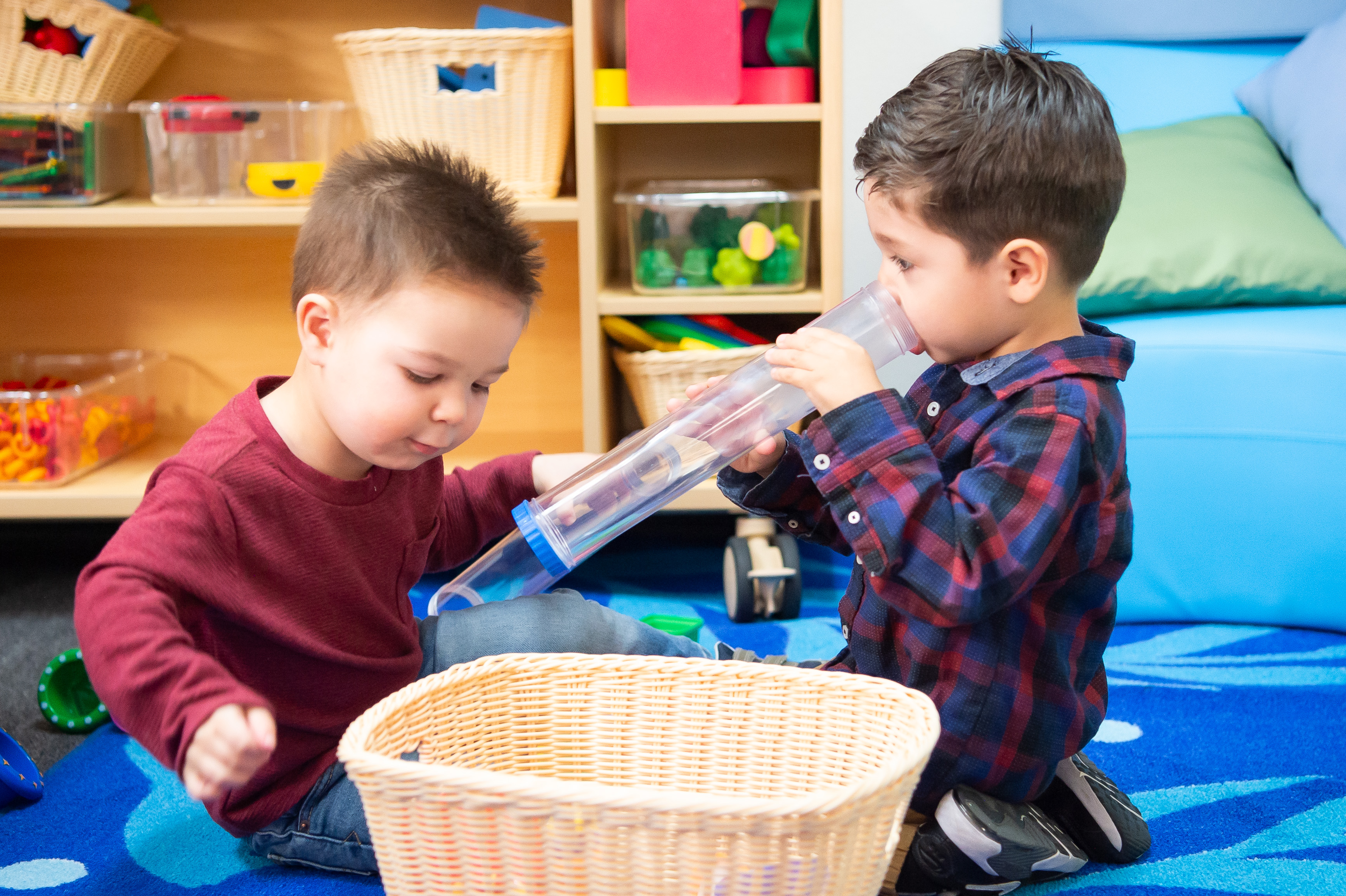 Dos niños pequeños, uno con una camisa roja y otro con una franela azul, juegan con juguetes en un aula colorida.