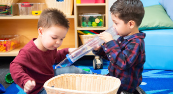 Dos niños pequeños, uno con una camisa roja y otro con una franela azul, juegan con juguetes en un aula colorida.