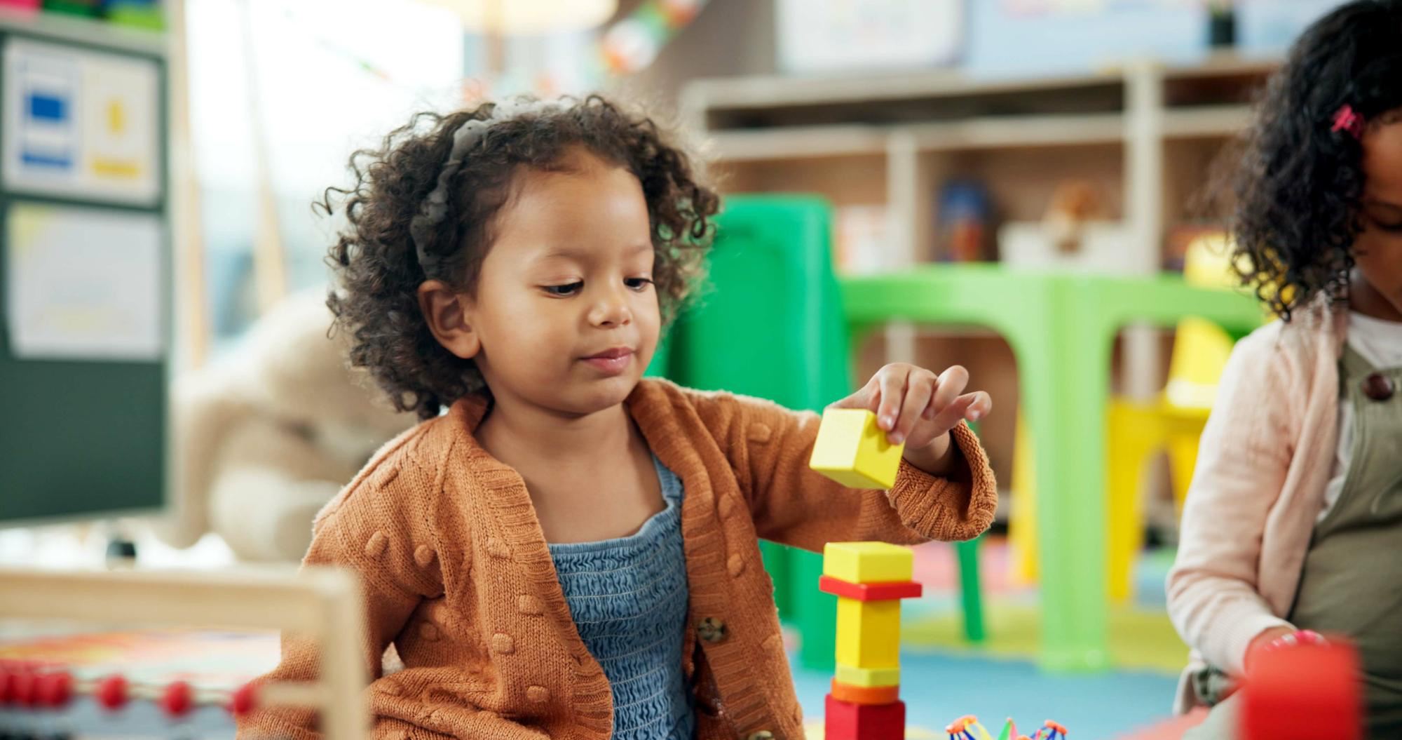 Una niña pequeña, en un entorno de aula, juega con bloques coloridos en el suelo, apilándolos y sonriendo.