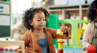 Una niña pequeña, en un entorno de aula, juega con bloques coloridos en el suelo, apilándolos y sonriendo.