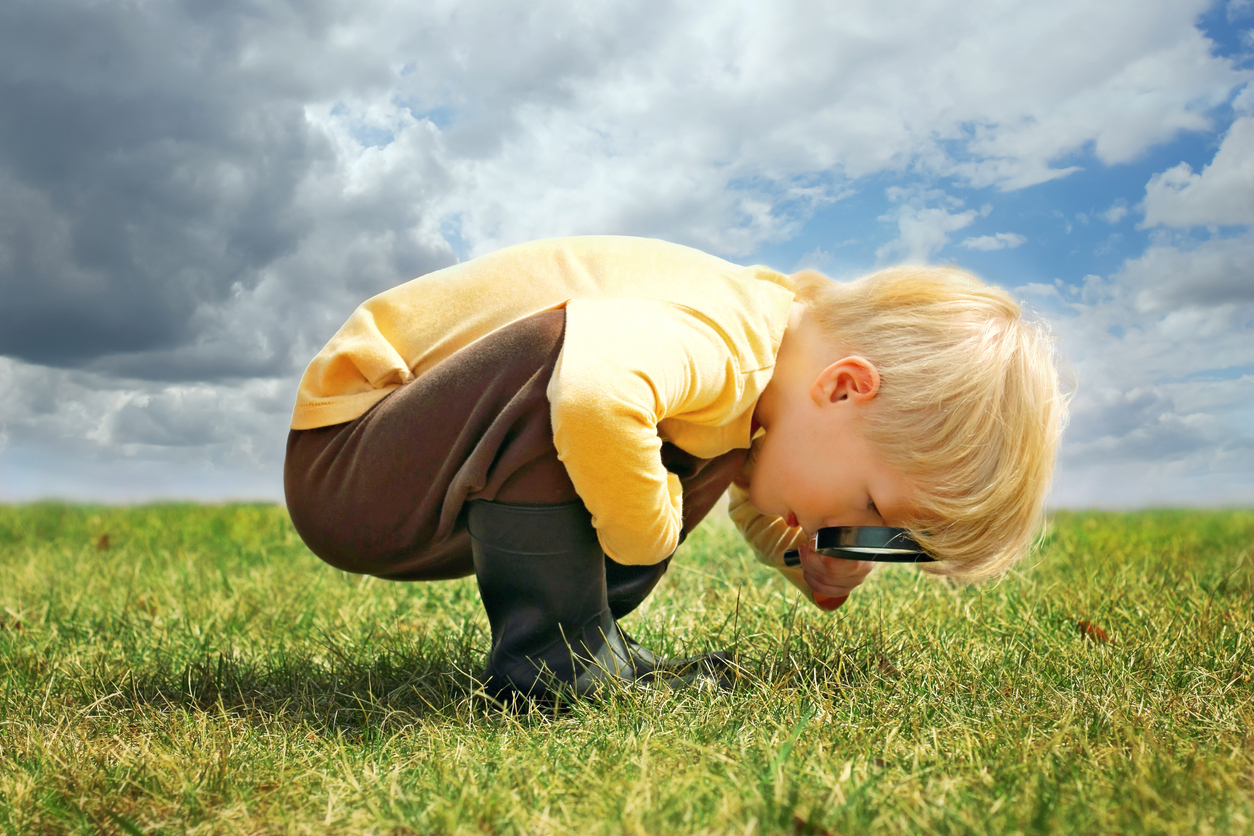 Niño pequeño con tez y pelo claros, al aire libre, agachado mirando al pasto a través de una lupa.