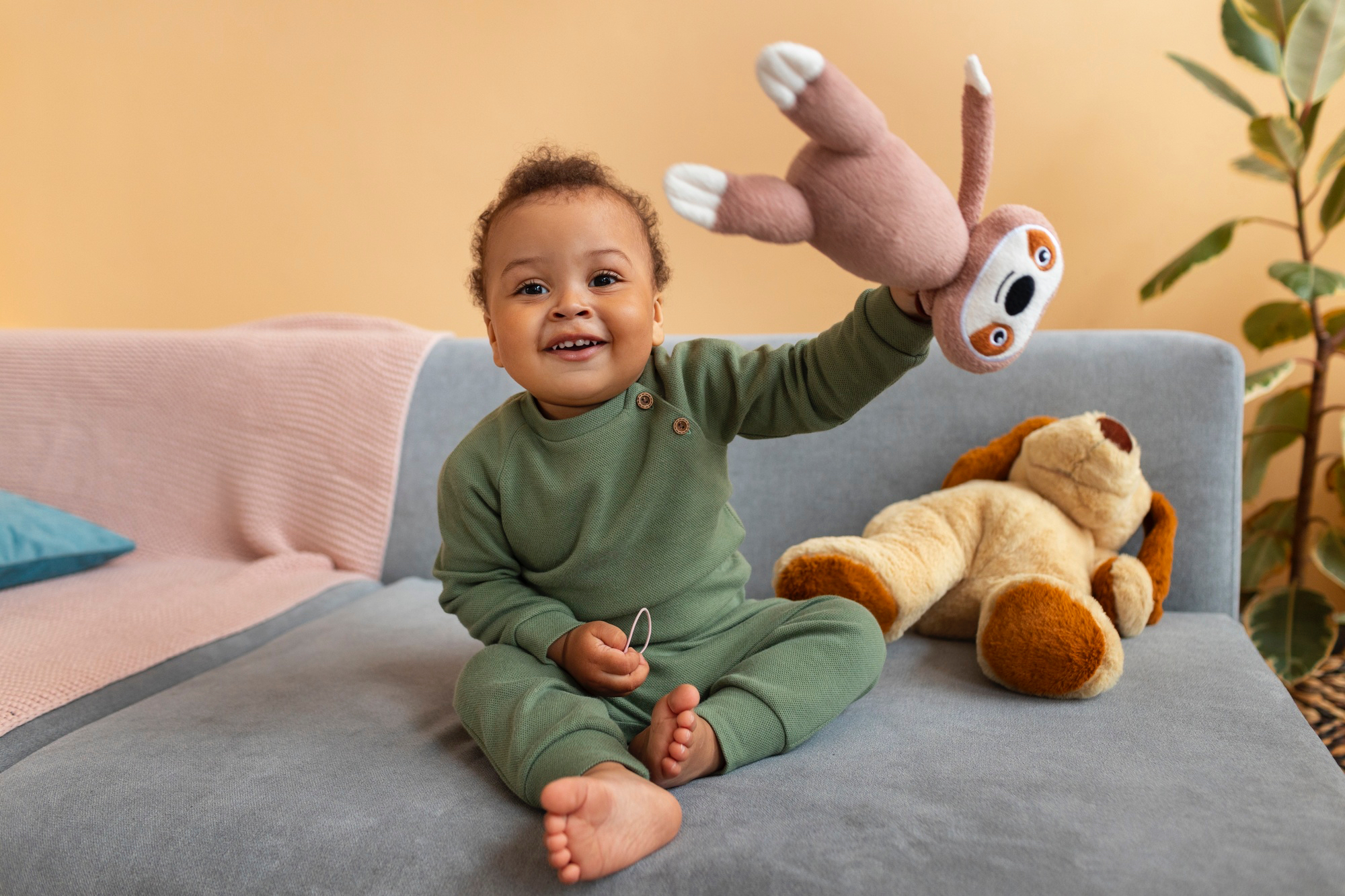 Un niño pequeño sonriente, vestido con un mameluco verde oscuro, está sentado en un sofá sosteniendo un peluche y sonriendo.