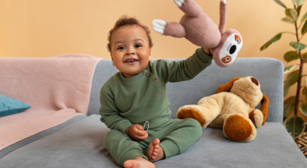 Un niño pequeño sonriente, vestido con un mameluco verde oscuro, está sentado en un sofá sosteniendo un peluche y sonriendo.