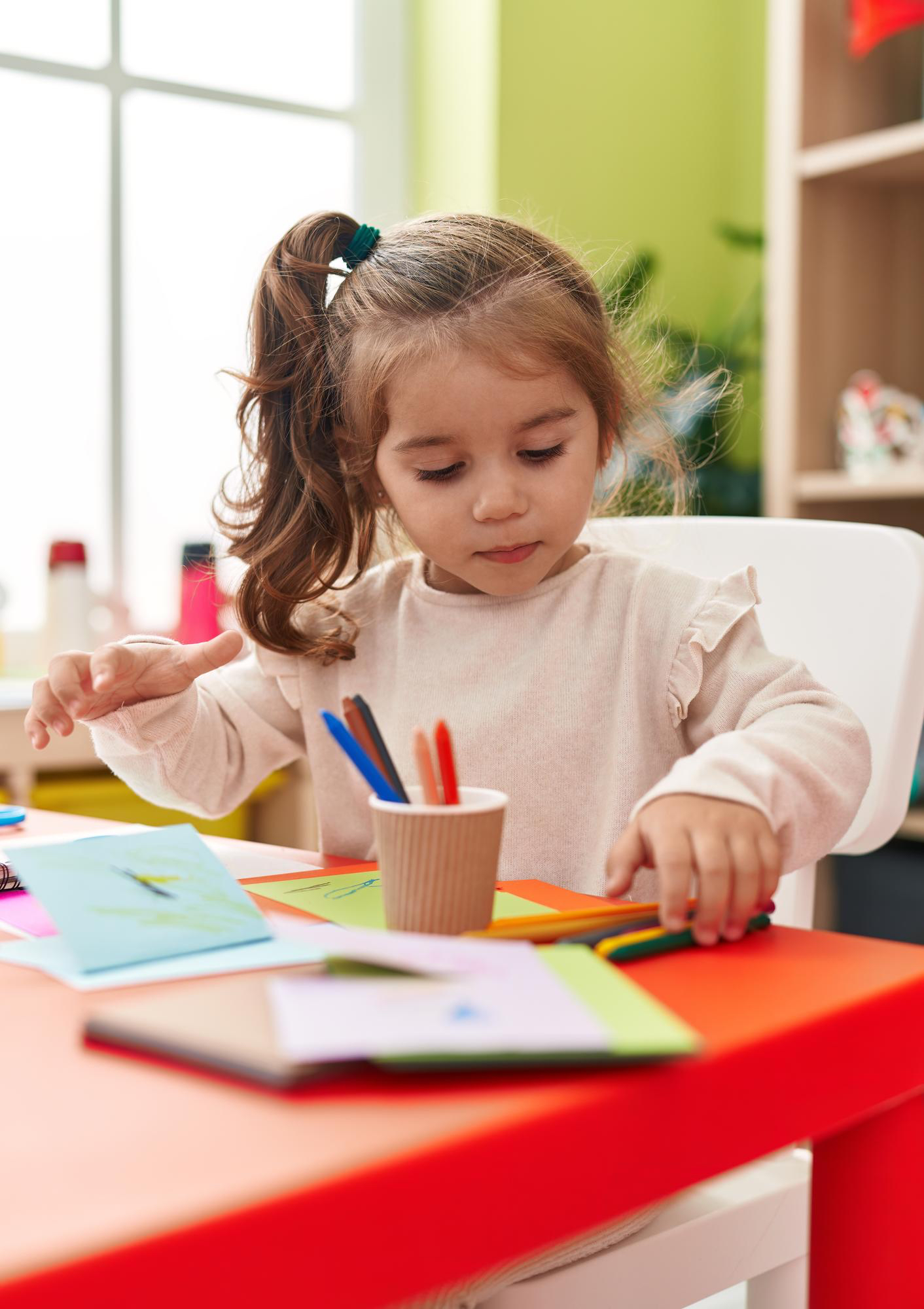 Niña pequeña sentada en un escritorio rojo en un aula, colorea una tarjeta con lápices de colores.