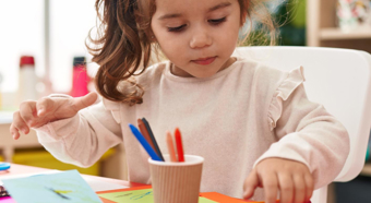 Niña pequeña sentada en un escritorio rojo en un aula, colorea una tarjeta con lápices de colores.