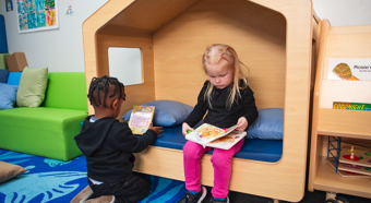 Dos niños pequeños sentados uno al lado del otro mientras leen libros infantiles en una casa de juegos para niños.