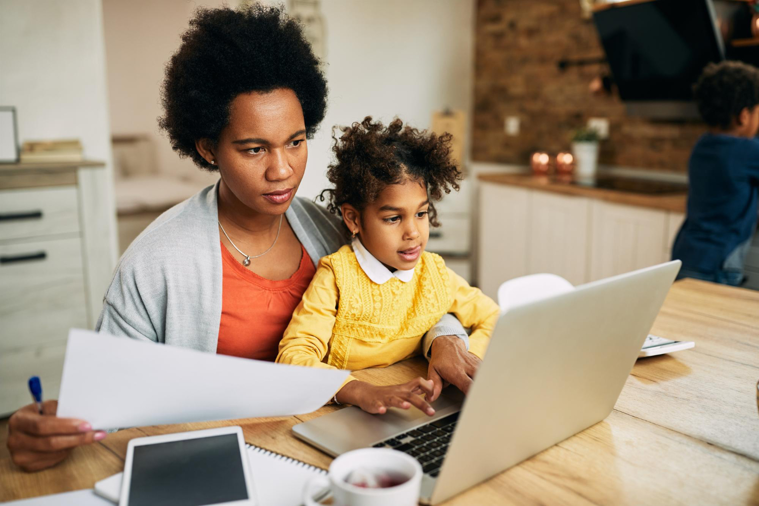 Una mujer adulta sentada a una mesa con un niño pequeño en su regazo, mientras mira la pantalla de una computadora y unos papeles que sostiene en las manos.