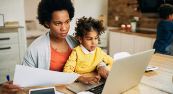 Una mujer adulta sentada a una mesa con un niño pequeño en su regazo, mientras mira la pantalla de una computadora y unos papeles que sostiene en las manos.