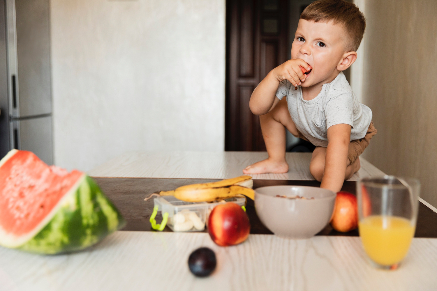 Un niño pequeño come una fruta en la cocina de su casa. También hay una sandía, un plátano, una manzana, jugo de naranja y otras frutas en la mesa.