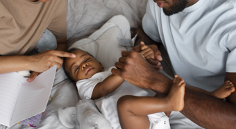 Dos figuras adultas se sientan en la cama con el bebé, ambas sonriendo y consolando al bebé en un entorno cómodo.