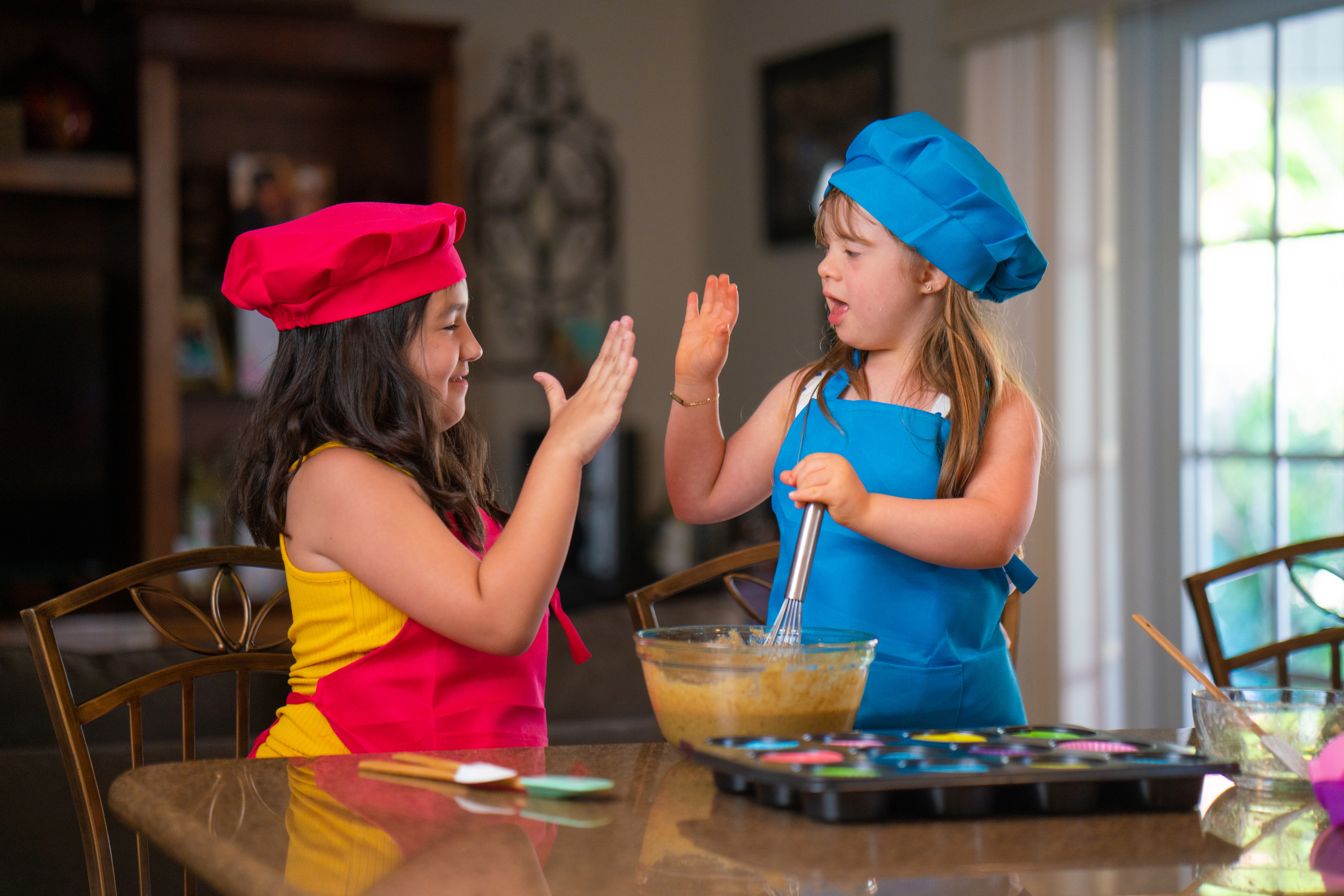 Dos niñas pequeñas de tez clara y cabello marrón, con trajes de cocineras, chocándose los cinco frente a una mesa con elementos de cocina.