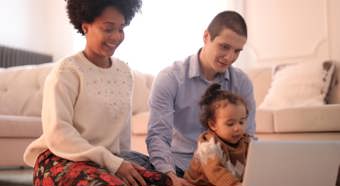 Una mujer y un hombre jóvenes se sientan con un niño pequeño en el suelo, mientras todos miran la pantalla de una computadora portátil. Un ambiente acogedor en el interior.