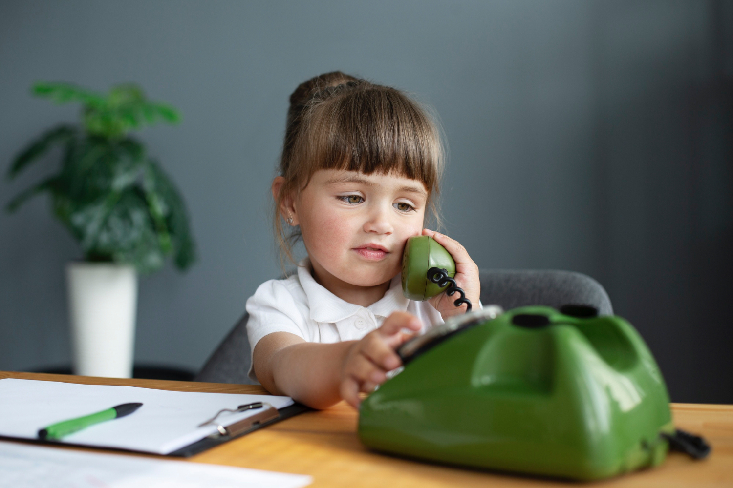 Una niña pequeña vestida con ropa bonita pretendiendo hablar por un teléfono verde, con papeles en el escritorio a su lado y una planta al fondo.