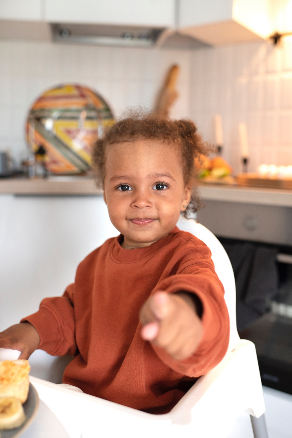 Una niña feliz sonríe y señala a la cámara. En una cocina interior, sentada en su trona.