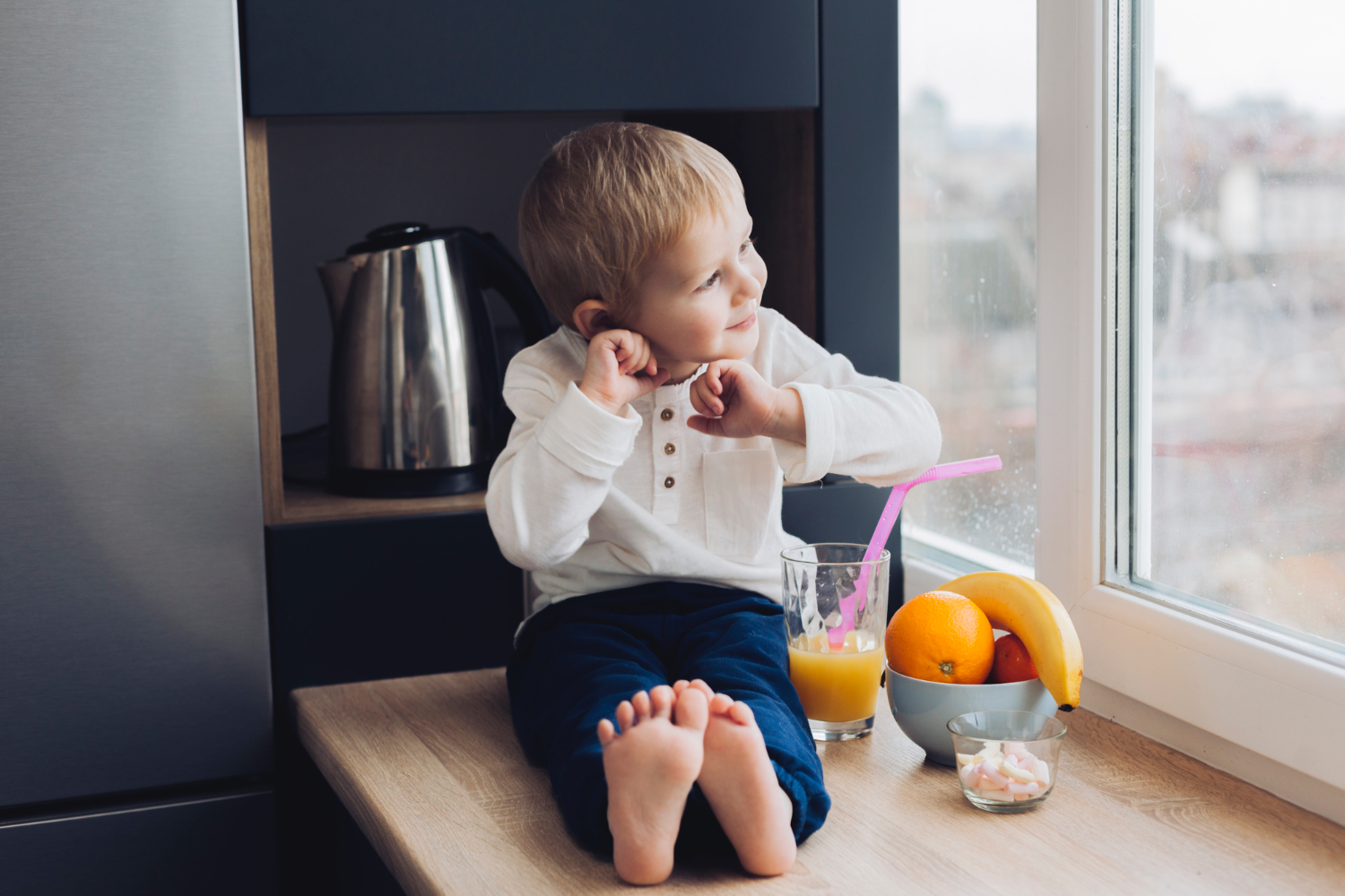 Un niño pequeño está sentado en el alféizar de la ventana, sonriendo y mirando hacia afuera. A su lado hay un vaso de jugo de naranja con una pajita rosa y un tazón de fruta.