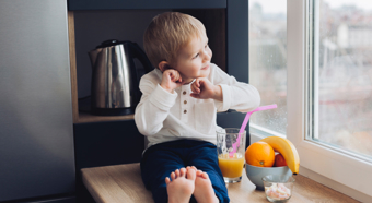 Un niño pequeño está sentado en el alféizar de la ventana, sonriendo y mirando hacia afuera. A su lado hay un vaso de jugo de naranja con una pajita rosa y un tazón de fruta.