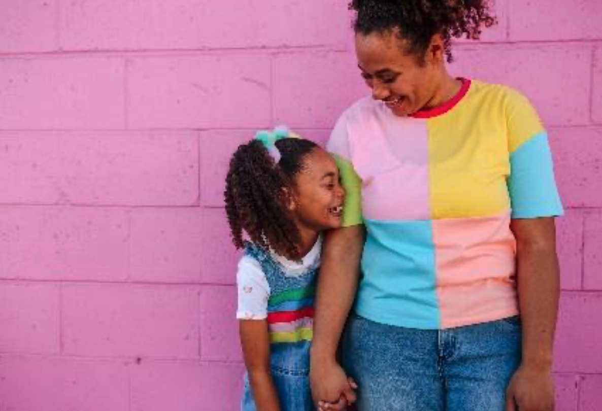 Una mujer adulta tomada de la mano con una niña riendo y sonriendo una a la otra, paradas frente a una pared rosa, ambas vistiendo ropa colorida.