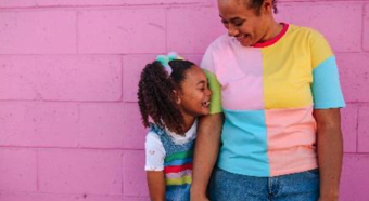Una mujer adulta tomada de la mano con una niña riendo y sonriendo una a la otra, paradas frente a una pared rosa, ambas vistiendo ropa colorida.