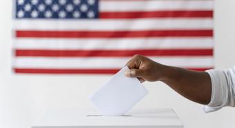 Mano emitiendo su voto en una urna, con la bandera estadounidense de fondo