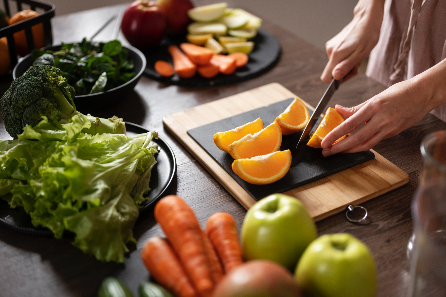 En una cocina donde una persona corta diversas frutas y verduras: lechuga, zanahoria, naranja, manzana y brócoli.