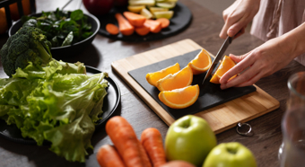 En una cocina donde una persona corta diversas frutas y verduras: lechuga, zanahoria, naranja, manzana y brócoli.