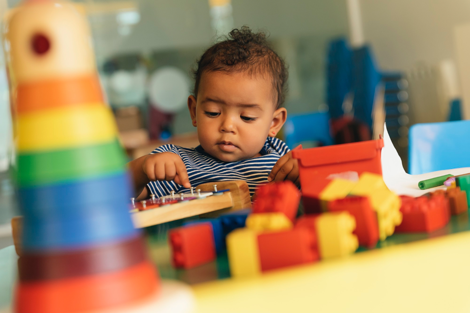 Un niño pequeño sentado a una mesa en un entorno de juego colorido, con bloques y otros juguetes sobre la mesa.