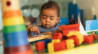 Un niño pequeño sentado a una mesa en un entorno de juego colorido, con bloques y otros juguetes sobre la mesa.