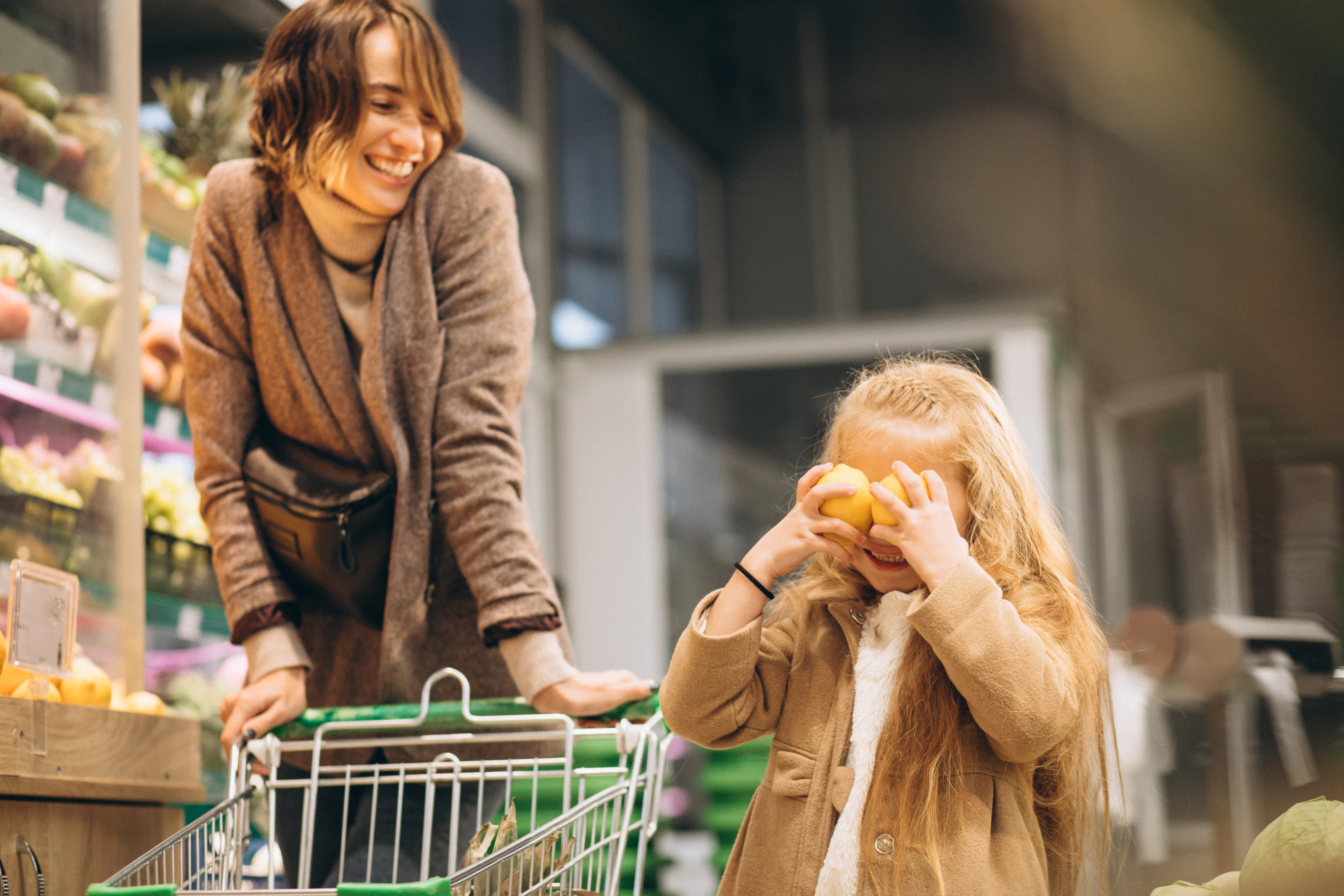 Mujer adulta llevando un carro de compras en un mercado, junto a una niña pequeña sonriente sujetando dos limones a la altura de sus ojos.