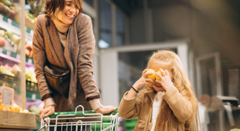 Mujer adulta llevando un carro de compras en un mercado, junto a una niña pequeña sonriente sujetando dos limones a la altura de sus ojos.