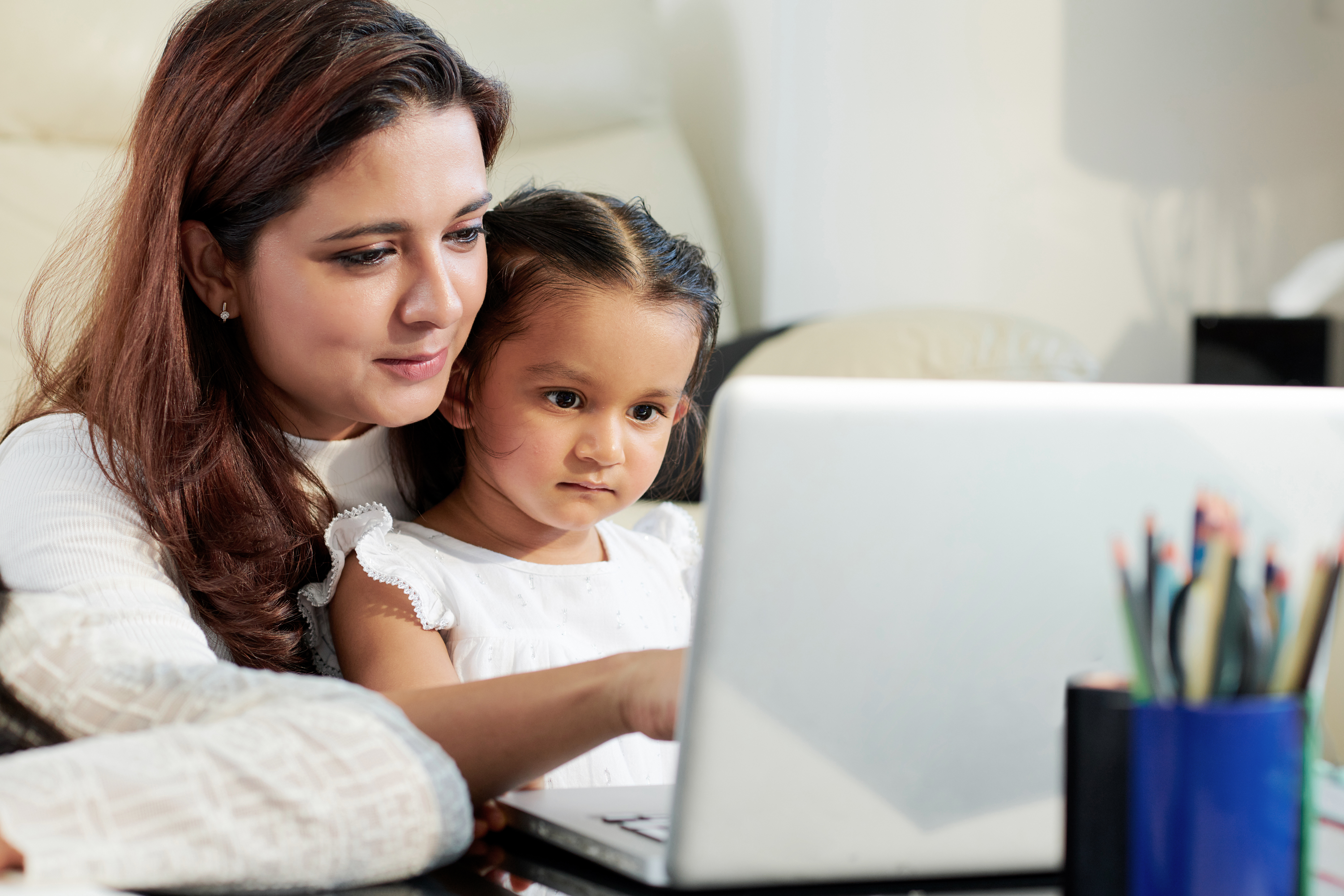 Mujer adulta y niña pequeña, de tez marrón clara y cabello castaño oscuro, mirando la pantalla de una computadora.