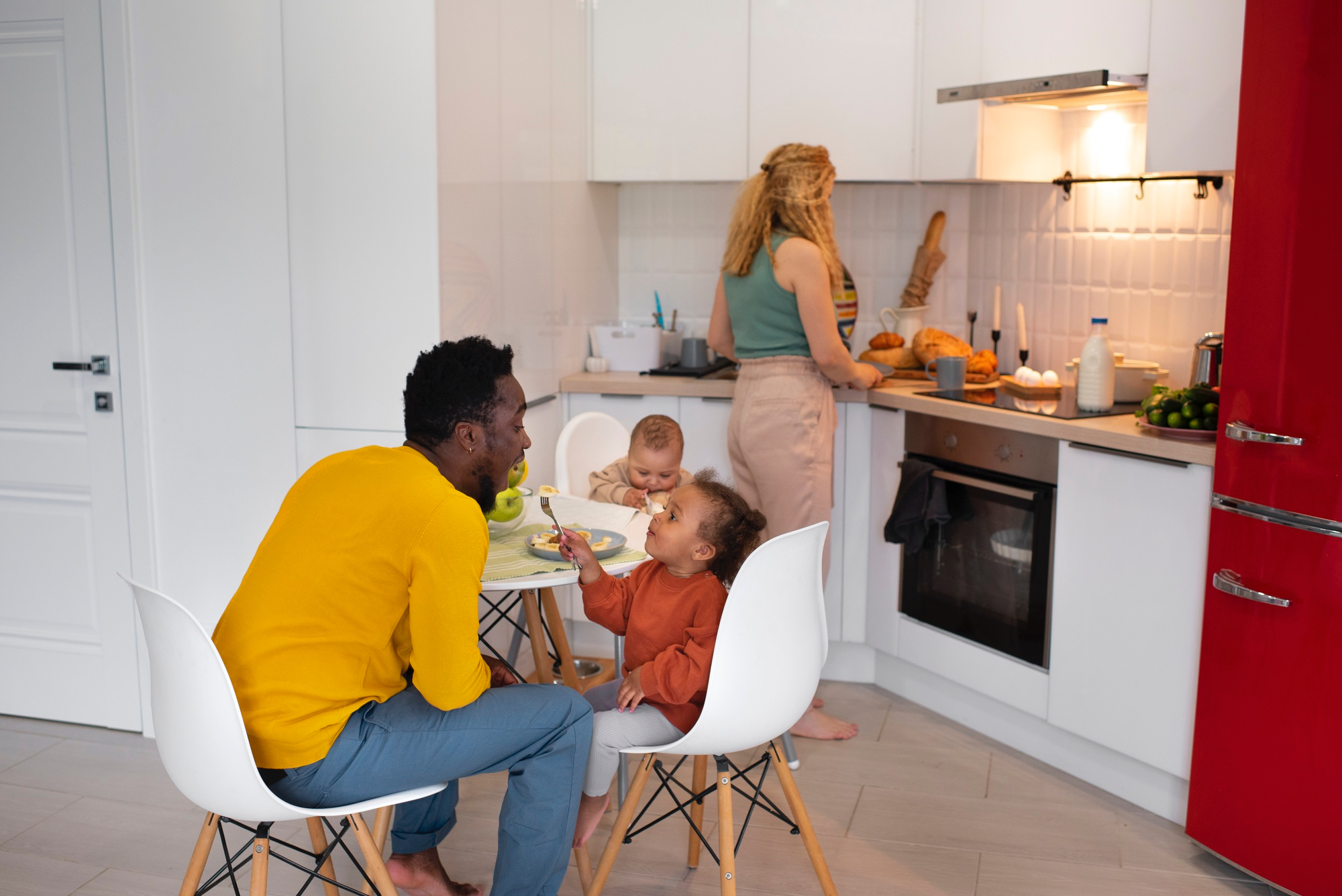 Una mujer y un hombre adultos en una cocina, con dos niños pequeños: uno sentado a la mesa con un tenedor en la mano y el otro en una silla alta. La mujer está cocinando.