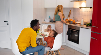 Una mujer y un hombre adultos en una cocina, con dos niños pequeños: uno sentado a la mesa con un tenedor en la mano y el otro en una silla alta. La mujer está cocinando.
