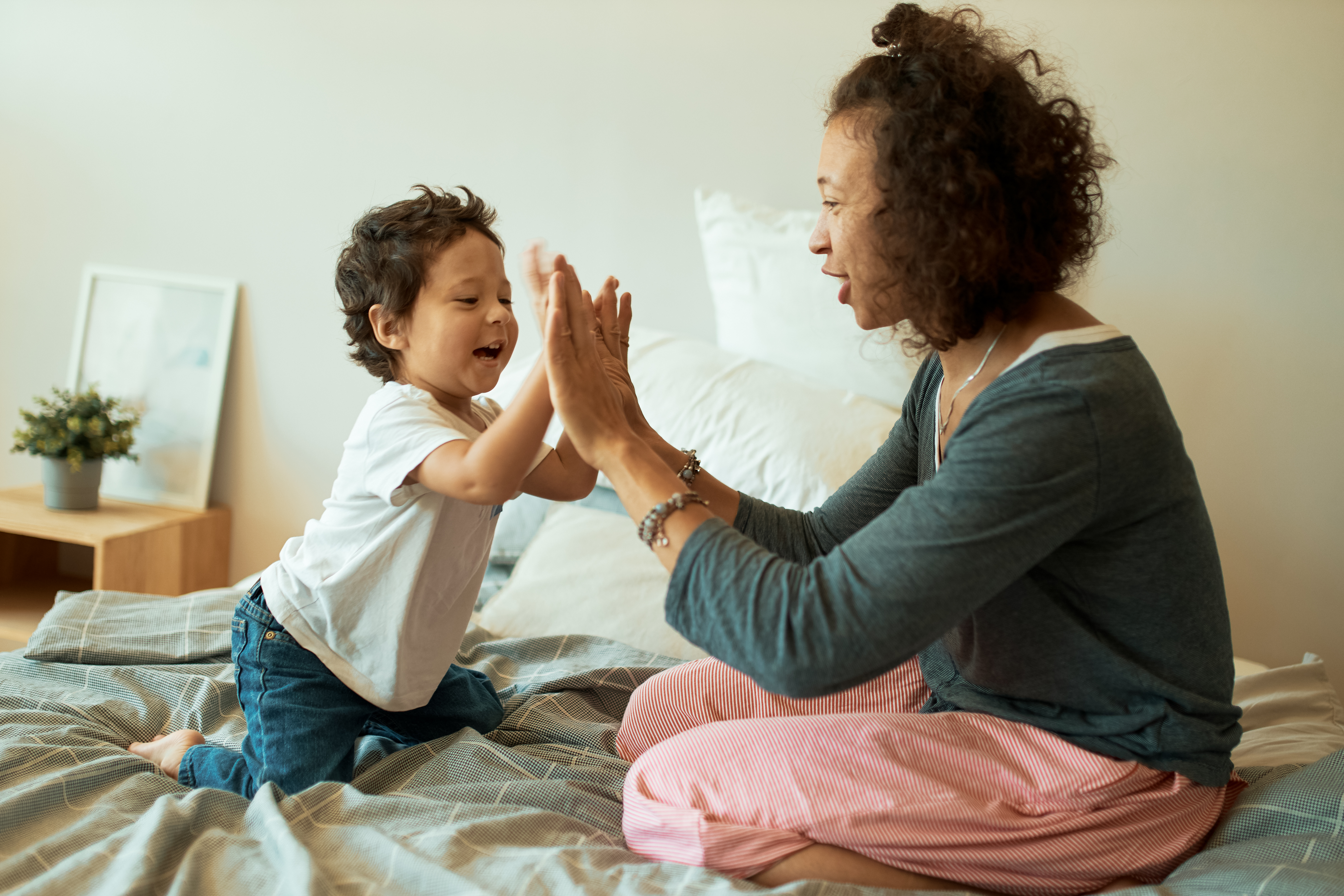 Mujeres felices con un niño jugando en casa, alegre mujer latina juntando las manos con un bebé sentado en la cama.