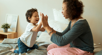 Mujeres felices con un niño jugando en casa, alegre mujer latina juntando las manos con un bebé sentado en la cama.