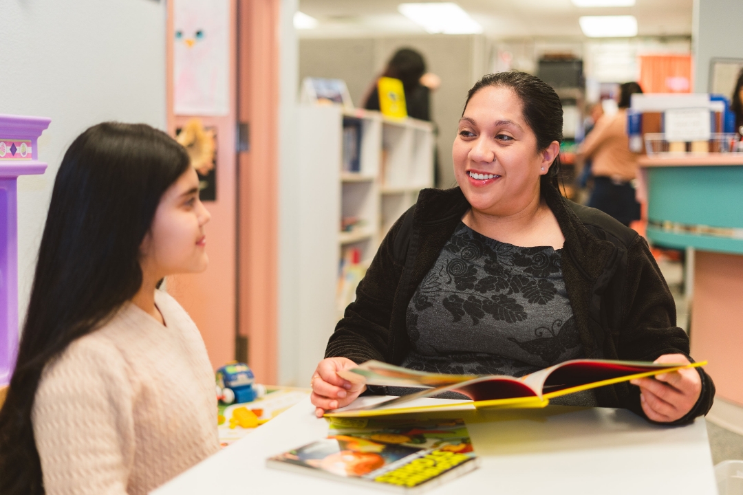 María Aguayo Galván en un salón de clases, sonriendo y hablando con una niña mientras sostiene un libro