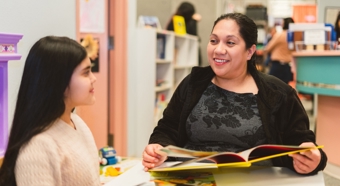 María Aguayo Galván en un salón de clases, sonriendo y hablando con una niña mientras sostiene un libro