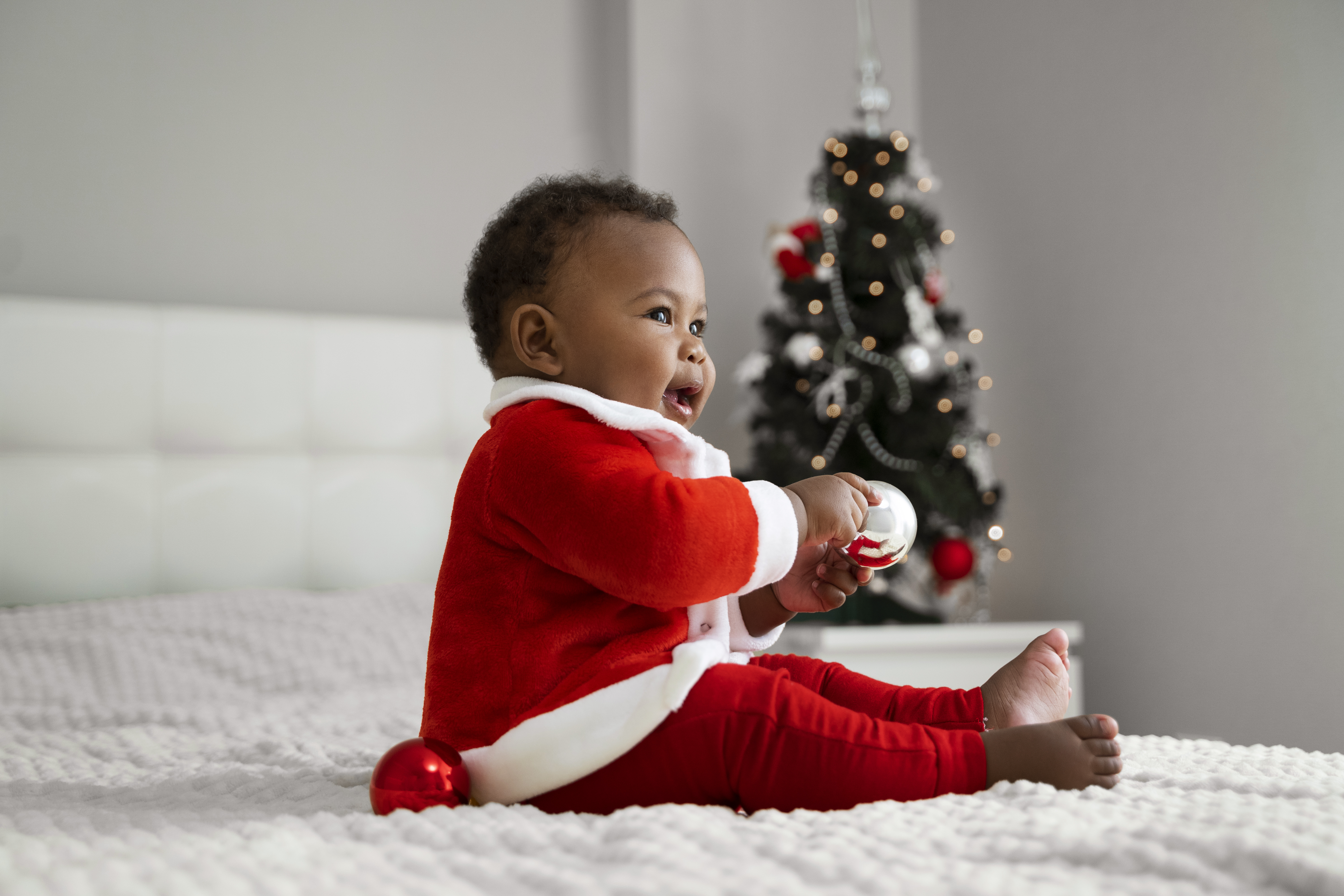 Vista lateral de un bebé sonriente en el interior con traje de Papá Noel, una cómoda cama blanca con un árbol de Navidad de fondo