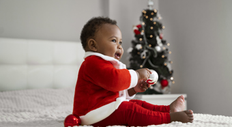 Vista lateral de un bebé sonriente en el interior con traje de Papá Noel, una cómoda cama blanca con un árbol de Navidad de fondo