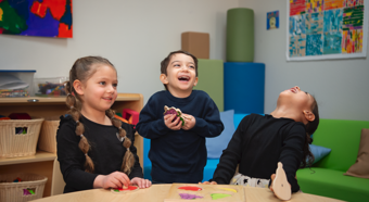 Dos niñas y un niño en un aula riéndose mientras juegan con juguetes